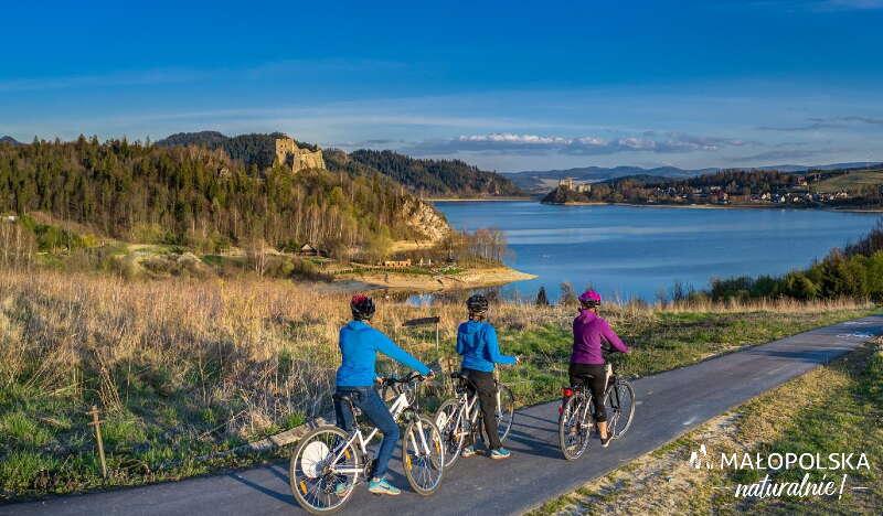 Trzy osoby na rowerach podziwiające widok na jezioro i zamek.