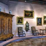 Komnata zamkowa, na ścianach wiszą obrazy, pod ścianami stoją od prawej drewniana komoda, stolik, dwa fotele, między którymi wisi zegar oraz duża ozdobna szafa, a na niej dwa dzbanki.