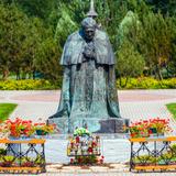 Pomnik - figura klęczącego przodem papieża świętego Jana Pawła II. Przed pomnikiem dwa klęczniki, kwiaty i znicze. Na drugim planie ławki, drzewa i trawnik.