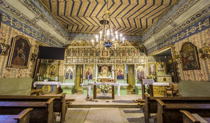 Wnętrze drewnianej cerkwi. Na ścianach polichromie i ikony, w nawie ławki, na wprost ołtarz, za nim piękny ikonostas.