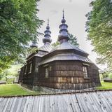 Drewniana cerkiew z zewnątrz, otoczona drewnianym ogrodzeniem. Wokół świątyni trawnik oraz kilka drzew.