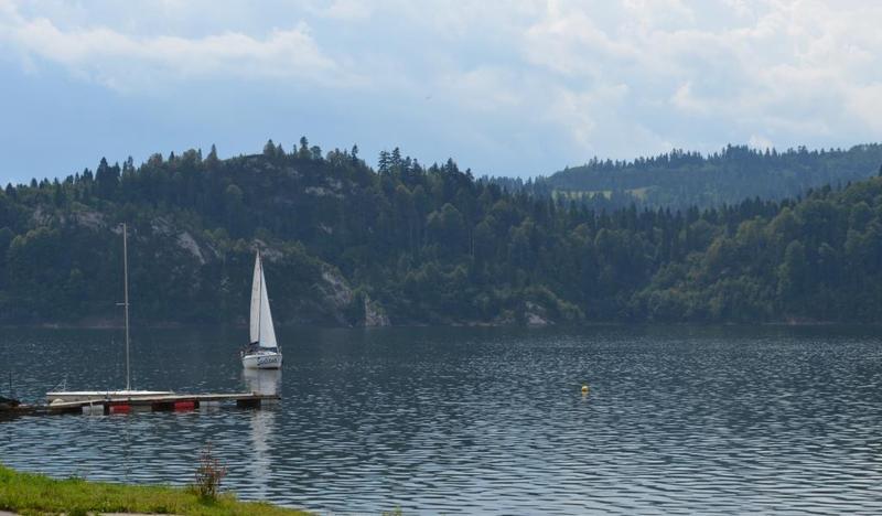 jezioro, na drugim planie zalesiona wzgórza, pływająca żaglówka i niewielki pomost po lewej