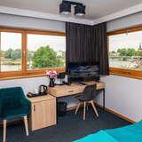 Pokój w hotelu, biurko, szafa, telewizor, dwa krzesła, widok przez okna na rzekę.