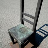 Zdjęcie krzesła od góry - rzeźby z oparciem i dwiema belkami w poprzek. Pod nim niski postument i wokół brukowany plac. Za nim padający jego cień.