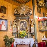 Wnętrze drewnianego kościoła z ołtarzem bocznym i amboną. Na drewnianej ścianie wisi obraz przedstawiający Jana Pawła II.