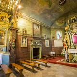 Wnętrze drewnianego kościoła - część prezbiterium z ołtarzem bocznym i ołtarzem głównym z krucyfiksem w centralnym punkcie. Na suficie i ścianach polichromia, wiszą również obrazy.