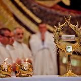 Na białym obrusie relikwie w monstrancji, w postaci fiolki z krwią św. Jana Pawła II. Po lewej dwie niskie, złote świece. W tle rozmyte trzy postacie kapłanów.