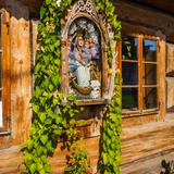 Kapliczka z figurą Matki Bożej z dzieciątkiem między oknami na drewnianym domu z bali, ozdobiona liśćmi i kwiatami.