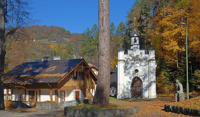 Mały jasny budynek murowanej kaplicy w Parku Zdrojowym. Po lewej willa w stylu szwajcarskim - widać dach i górne piętro.