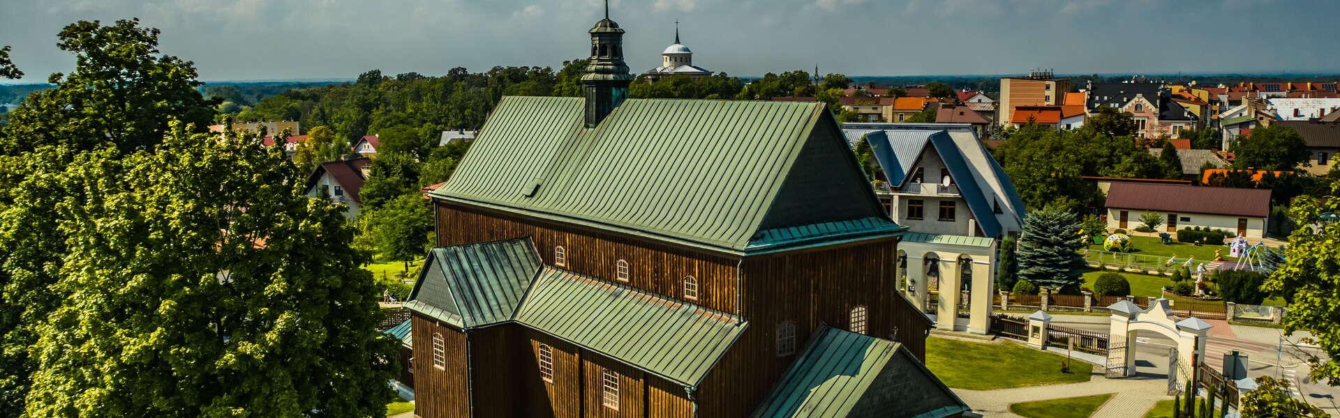 Widok na drewniany kościół w Dąbrowie Tarnowskiej. Wokół budynku znajdują się drzewa. W tle zabudowania miasta.
