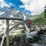 Turysta z przewodnikiem w Tatrach przechodzący przez drewniany mostek. W tle ośnieżone góry Tatry.