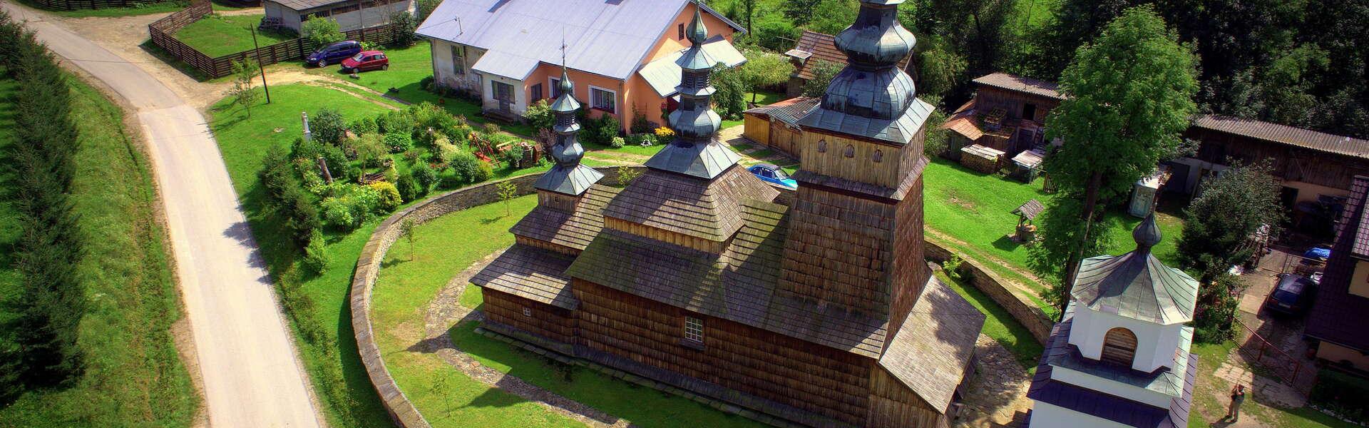 Widok na bryłkę drewnianej cerkwi z lotu ptaka. Trzy kopuły ze strzelistymi wieżami. Wokół cerkwi ogrodzenie. W tle zabudowania domów.