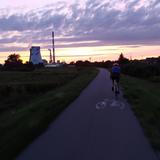 Zmrok, zachód słońca. Po wiślanej Trasie Rowerowej jedzie rowerzysta, po bokach ciemne krzewy. W oddali widać wysokie kominy. Niebo częściowo zachmurzone.