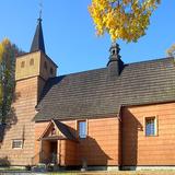 Drewniany kościół z wieżą i znajdującą się pośrodku dachu wieżyczką na sygnaturkę. Wokół kilka drzew w jesiennych kolorach.