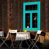 Pod drewnianą ściana z oknem o błękitnych framugach, stoją białe metalowe stoliki i krzesła przykryte koronkowymi obrusami.