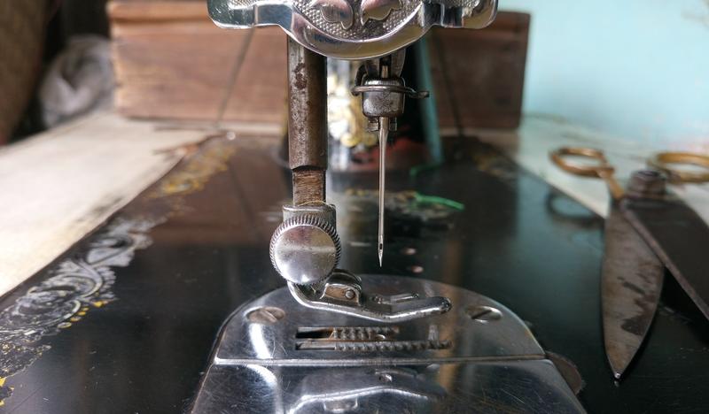 Zdjęcie ukazujące na pierwszym planie igłę oraz stopkę starej maszyny do szycia. W tle widoczne stare nożyce.