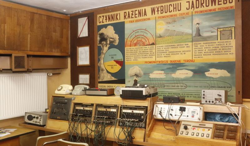 Aparatura telekomunikacyjna, telefony, radiostacje w sekcji alarmowania. Na ścianach tablica informacyjna o wybuchu jądrowym.
