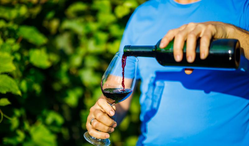 Zdjęcie przedstawia mężczyznę nalewającym czerwone wino z winnicy zawisza do kieliszka. W tle widoczna rozmyte krzewy winorośli.