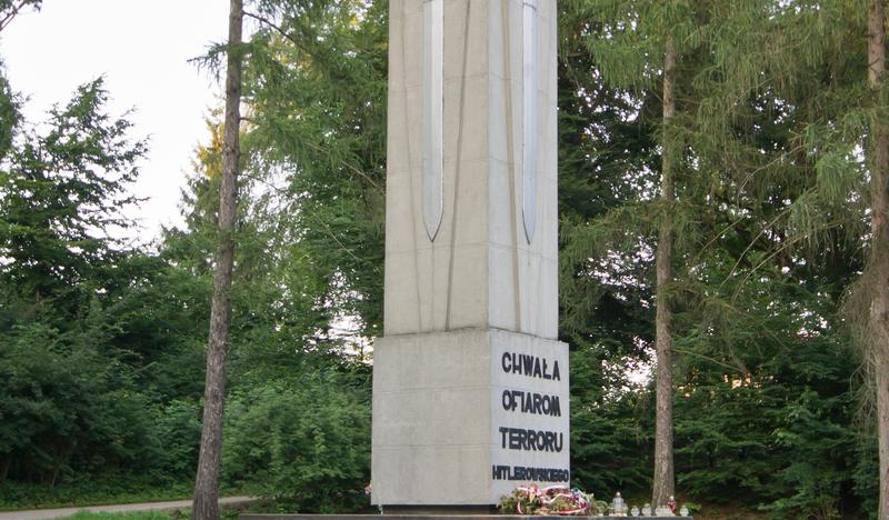 Fragment lasu. W nim Obelisk ze zniczem na szczycie i tablicą pamiątkową.