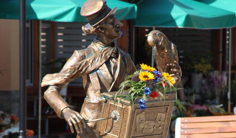 Pomnik mężczyzny z katarynką, na której siedzi papuga i leżą kwiaty.