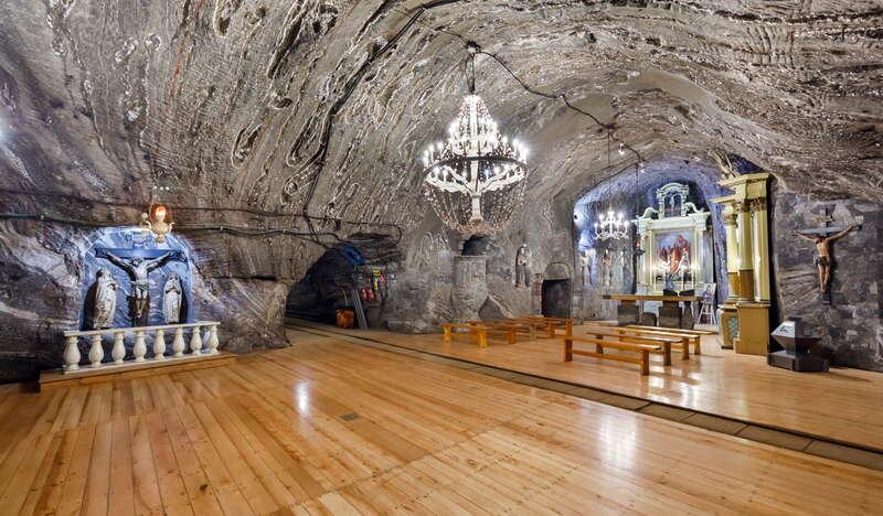 Widok na podziemną kaplicę świętej Kingi w Kopalni soli Bochnia. Z lewej krzyż wiszący na ścianie, po prawej ołtarz a przed nim ławki. Po środku kryształy żyrandol.