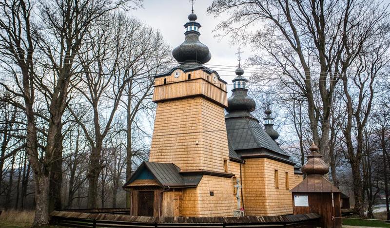 Jasna drewniana cerkiew z wysoką wieżą. Na dachach kopuły pokryte blachą. Zdjęcie późnojesienne z bezlistnymi drzewami.