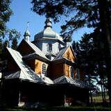 Drewniana cerkiew wśród drzew. Oświetlony dach i kopuły, reszta budynku w cieniu.
