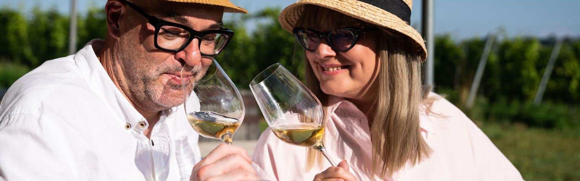 Kobieta i mężczyzna w jasnych koszulach i słomkowych kapeluszach rozkoszują się winem, w tle winnica.
