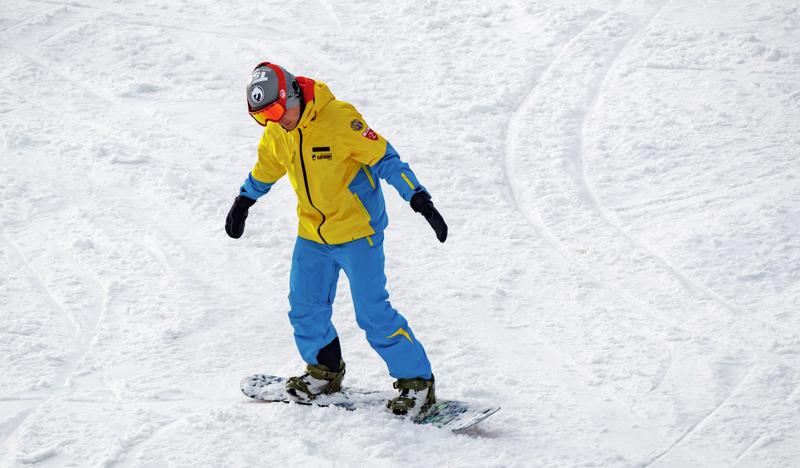 Mężczyzna w żółto-niebieskim komplecie sportowym zjeżdża na desce snowboardowej po ośnieżonym stoku