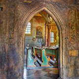 Wnętrze drewnianego kościoła - drewniany rzeźbiony portal, widać przez niego drewniane zdobione rzeźbieniami ławki dla wiernych w kolorze brązowo-zielonym. Nad nimi obraz na ścianie i po lewej małe okno. Po prawej złota kolumna i w głębi bocznego ołtarza małe okno.