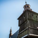 Górna część drewnianej wieży kościoła w ciemnym kolorze. Wieża zwieńczona blaszaną wieżyczką z krzyżem.