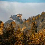Spowite mgłą ruiny murowanego  zamku na wzgórzu z wieżą kwadratową z otworami i z fragmentem murów obronnych. Niżej zamku na około lasy w jesiennym klimacie, ze złotymi liśćmi na drzewach. Ponad mgłą fragment pogodnego nieba.