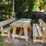 Drewniane ławki oraz ława w ogrodzie stojące w cieniu. Z tyłu kilka drzew.