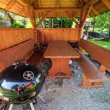 Drewniana wiata grillowa usytuowana w ogrodzie. Na pierwszym planie grill w kolorze czarnym, a z tyłu stół z dwoma ławkami.