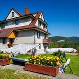 Duży biało brązowy dom Julia w Wysowej-Zdrój, przed nim zielony trawnik, donice z kwiatami i huśtawka, a z tyłu zielone wzgórza oraz niebieskie niebo.