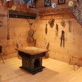 Drewniane pomieszczenie, na środku którego stoi drewniany zabytkowy stół a na ścianach wiszą zabytkowe eksponaty, w tym: skrzypce i inne instrumenty muzyczne, naczynia, chusta, obrazy.