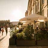 Zewnętrzny ogródek Restauracji Bianca w Krakowie z widokiem na Rynek Główny i Sukiennice. Wokół ogródka rosną rośliny w donicach, a nad stolikami znajdują się parasole z nazwą restauracji.