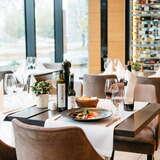 Widok na nakryte stoły w Restauracji Qualita w Krakowie. Na stole widocznym na pierwszym planie widać jedzenie na talerzu, wino i dodatki do posiłku. W oddali okna oraz półeczki na wina.