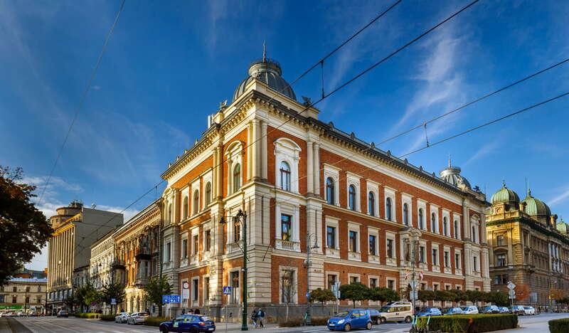 Eleweacja budynku Akademii Sztuk Pięknych w Krakowie. Budynek jest wykonany z czerwonej cegły oraz beżowych elementów. Na górze widać kopułę. Obok budynku są inne kamienice, po ulicy jadą auta, a nad nią są tramwajowe przewody trakcyjne