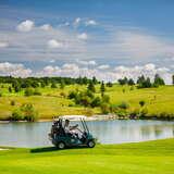 Niebieski wózek golfowy z kobietą i mężczyzną w środku jedzie po idealnie skoszonym, zielonym polu golfowym w Paczółtowicach. Za wózkiem znajduje się staw, a nieco dalej niskie drzewa, a nad nimi niebieskie niebo.