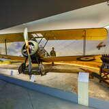 Mały samolot wojskowy w hangarze w Muzeum Lotnictwa w Krakowie. Obok niego stoją eksponaty wojenne, a z tyłu postać z kartonu.