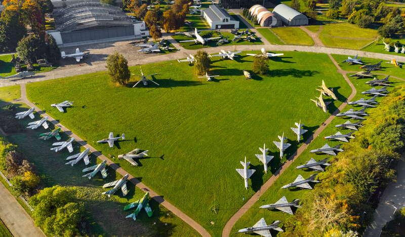 Duża, zielona przestrzeń z alejkami, wzdłuż których równo stoją samoloty. Na górze zdjęcia widać kilka budynków Muzeum Lotnictwa w Krakowie, w tym duży hangar.