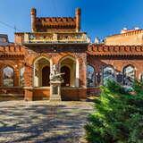 Widok na bryłę płaskiego, neogotyckiego budynku z cegły Muzeum Celestat w Krakowie. W oknach obrazy. Przed budynkiem rzeźba. Przed budynkiem brukowany plac. Po prawej duży krzew. U góry bezchmurne niebo.