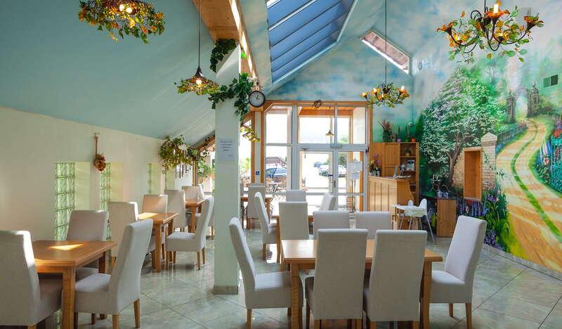 Jadalnia, stoły i białe tapicerowane krzesła z wysokim oparciem, na ścianach kolorowe malowidła.