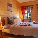 Wnętrze przytulnej sypialni, drewniane ściany, łóżko z beżową narzutą, okno z zasłonkami w czerwoną i granatową kratę