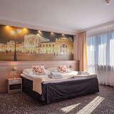 Pokój w pastelowych kolorach, dwuosobowe łóżko, duże okno z firanką i zasłonami.