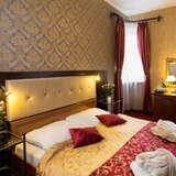 Łóżko dwuosobowe z kolorową narzutą, wzorzysta tapeta, okno z firanką, telewizor na ścianie.