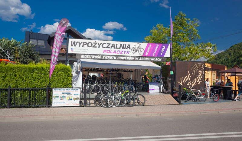Stoisko wypożyczalni rowerów tuż przy ulicy, charakterystycznych biało-różowo-fioletowy baner reklamowy wypożyczalni