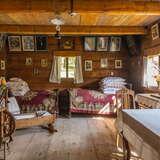 Wnętrze starej drewnianej, chłopskiej chaty, stół, dwa łóżka zasłane kolorowymi kapami, wiele poduszek, dużo obrazków w drewnianych ramach na ścianach, drewniany kołowrotek.