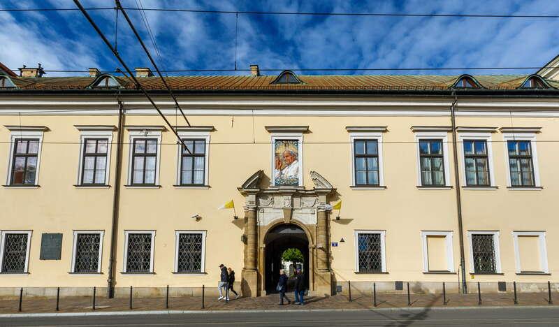 Beżowy jednopiętrowy budynek z Oknem Papieskim w Krakowie. Z bramy pod Oknem zwiewają dwie biało-żółte flagi. Przed budynkiem znajdują się tory tramwajowe, a nad nim niebieskie niebo.